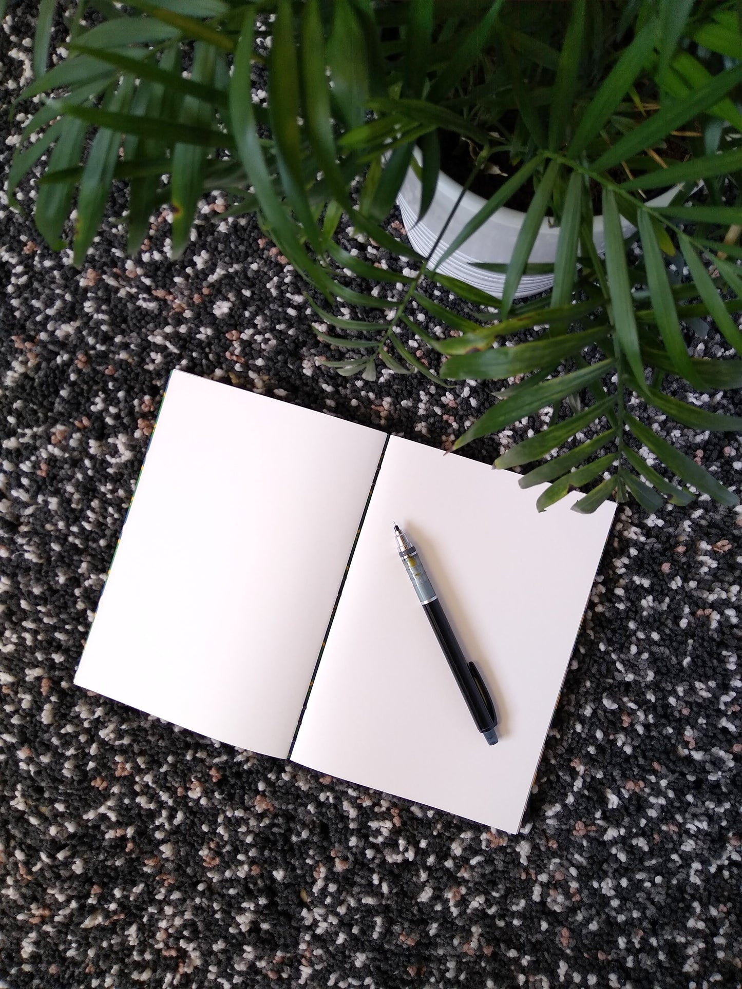 An open journal lays on a black shag carpet next to a potted plant. The journal has a black thread connecting the pages at the spine and a black mechanical pencil rests across the blank white pages.