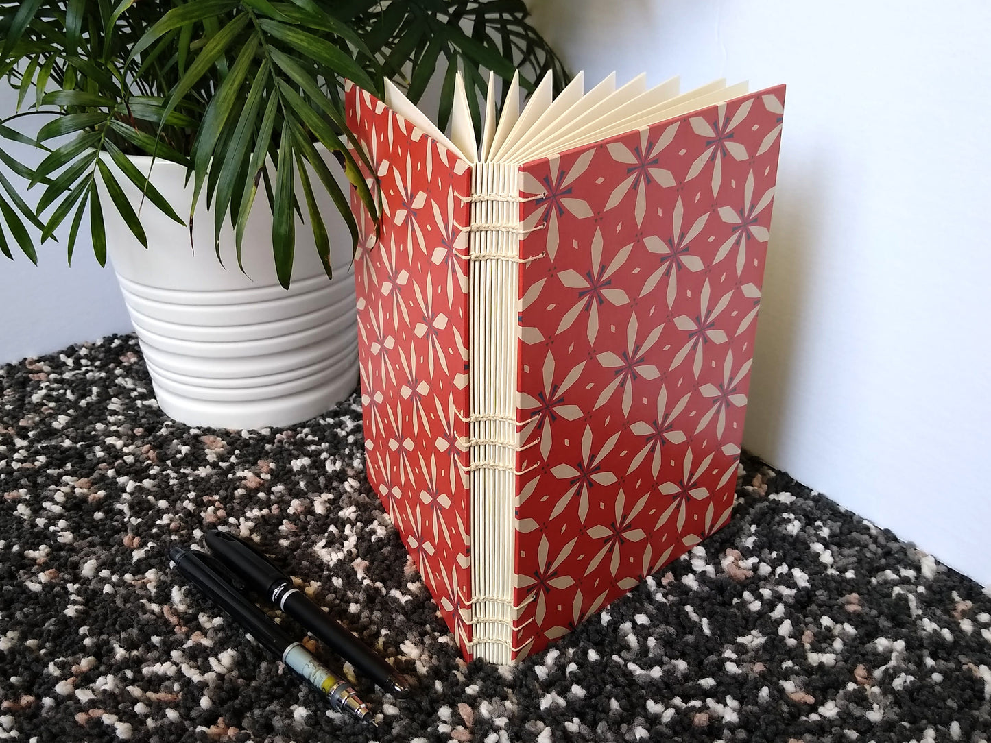 A handmade journal sits upright beside a potted plant, and a black pen and mechanical pencil. The cover of the journal has a red and cream geometric floral design on it. The open spine and cream thread stitching are visible.
