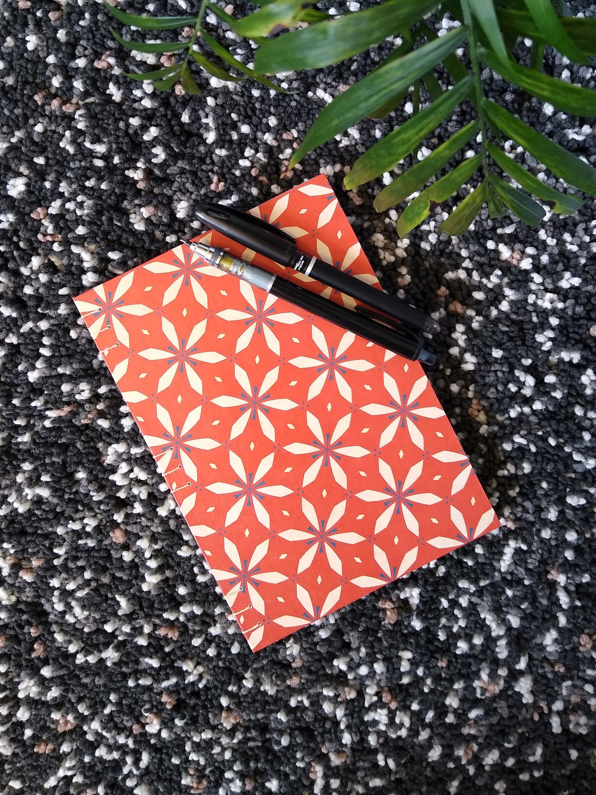 A handmade journal laying on a black shag carpet beside a potted plant, and with a black pen and mechanical pencil on top. The cover of the journal has a red and cream geometric floral design on it.