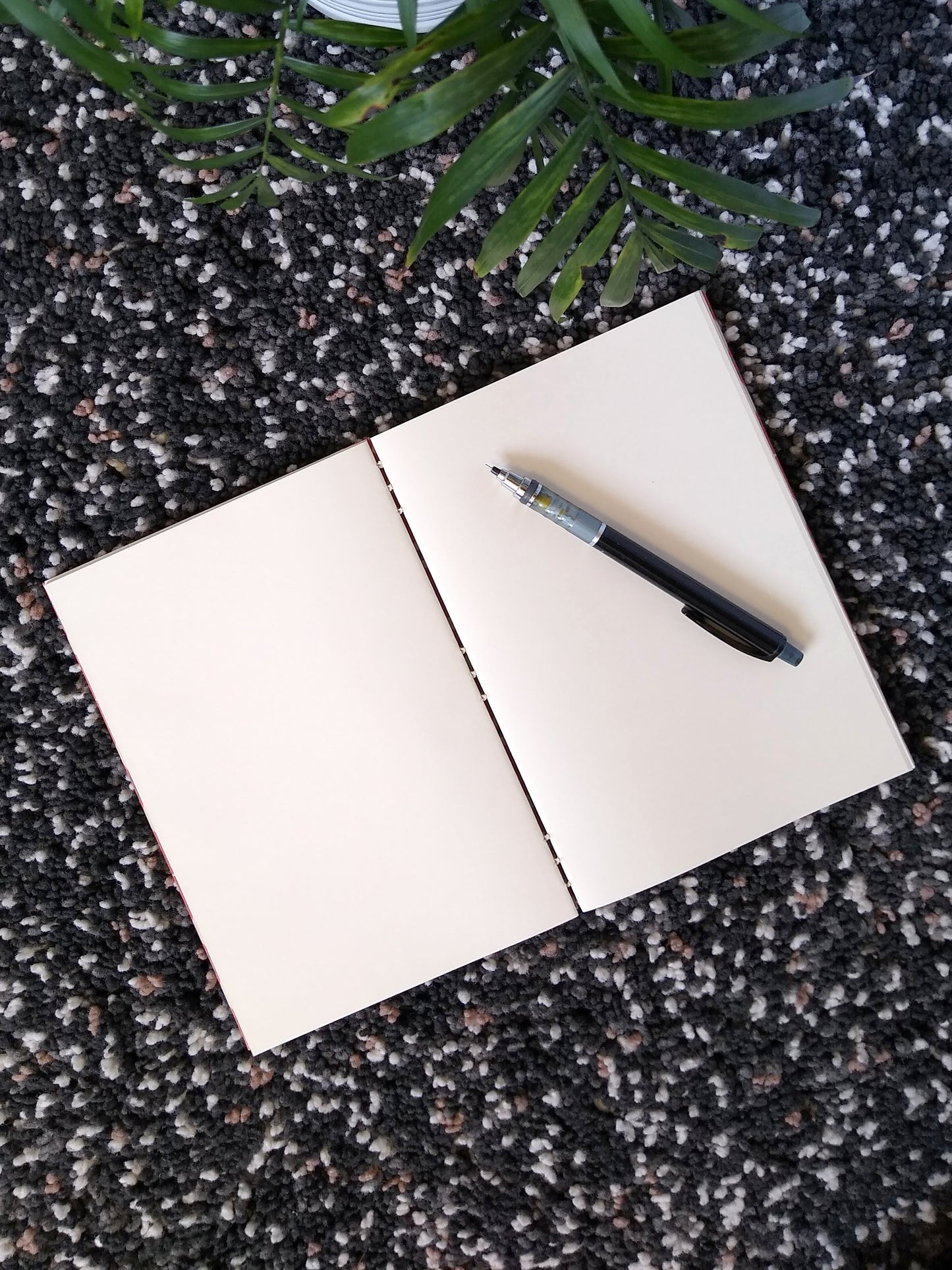 An open journal lays on a black shag carpet next to a potted plant. The journal has a cream thread connecting the pages at the spine and a black mechanical pencil rests across the blank cream pages.