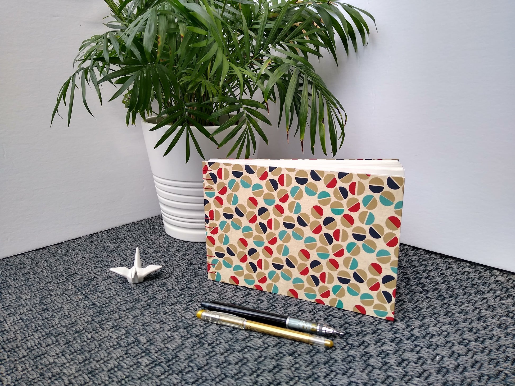 A handmade journal sits upright beside a potted plant, a ceramic crane, a gold pen, and a black mechanical pencil. The cover of the journal is cream with half circles in gold, teal, dark blue, and red all over it. The half circles are placed next to each other to look like the head of a flat-head screw.