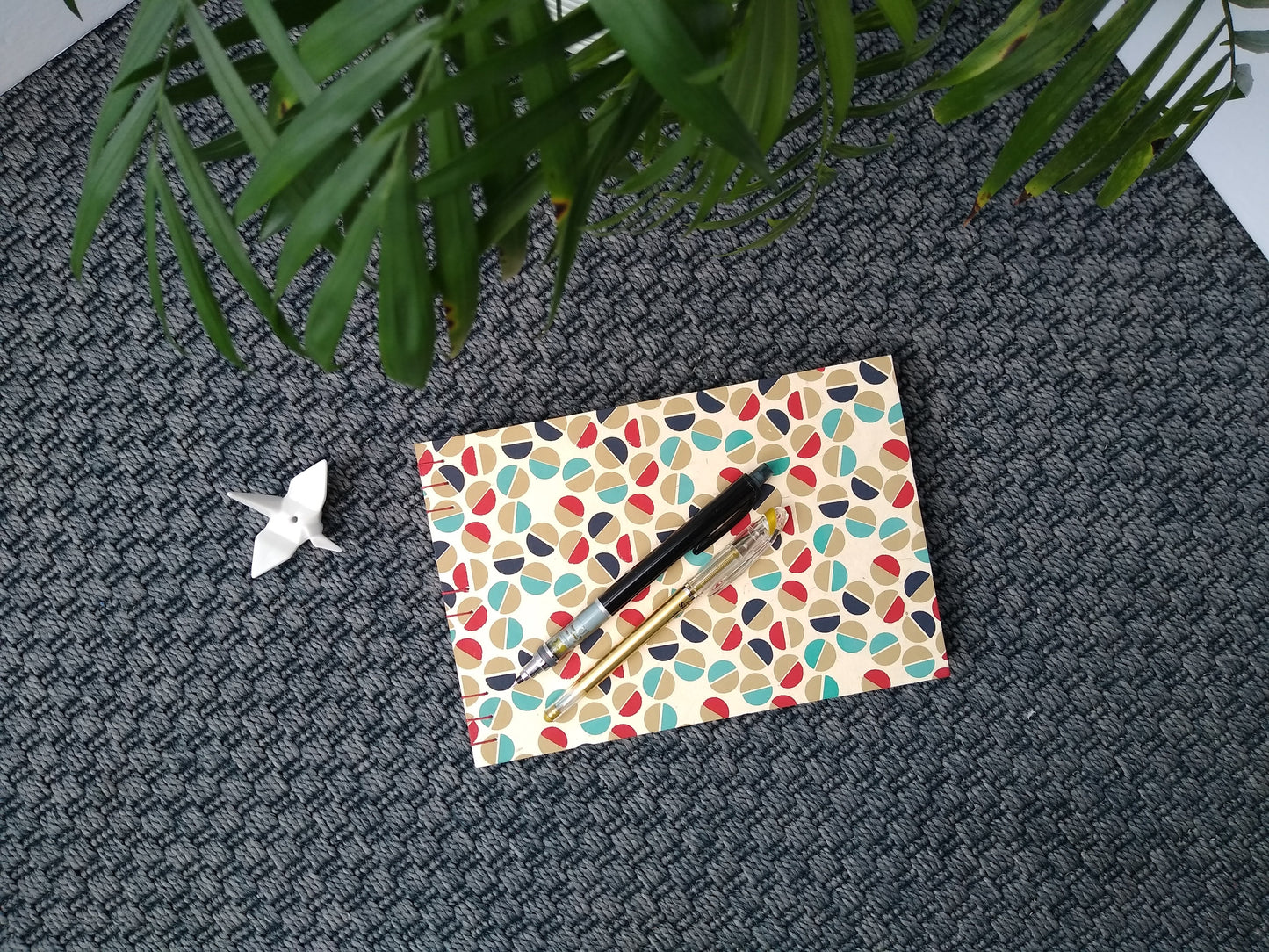 A handmade journal laying on a grey carpet beside a potted plant, a ceramic crane, a gold pen, and a black mechanical pencil. The cover of the journal is cream with half circles in gold, teal, dark blue, and red all over it. The half circles are placed next to each other to look like the head of a flat-head screw.