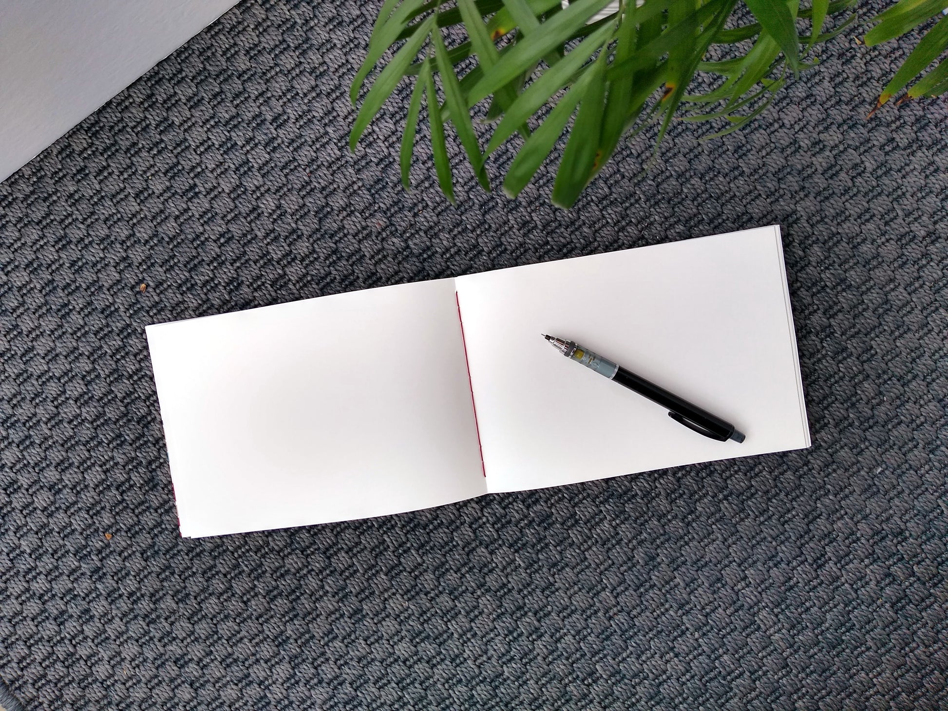 An open journal lays on a grey carpet next to a potted plant. The journal has a red thread connecting the pages at the spine and a black mechanical pencil rests across the blank white pages.