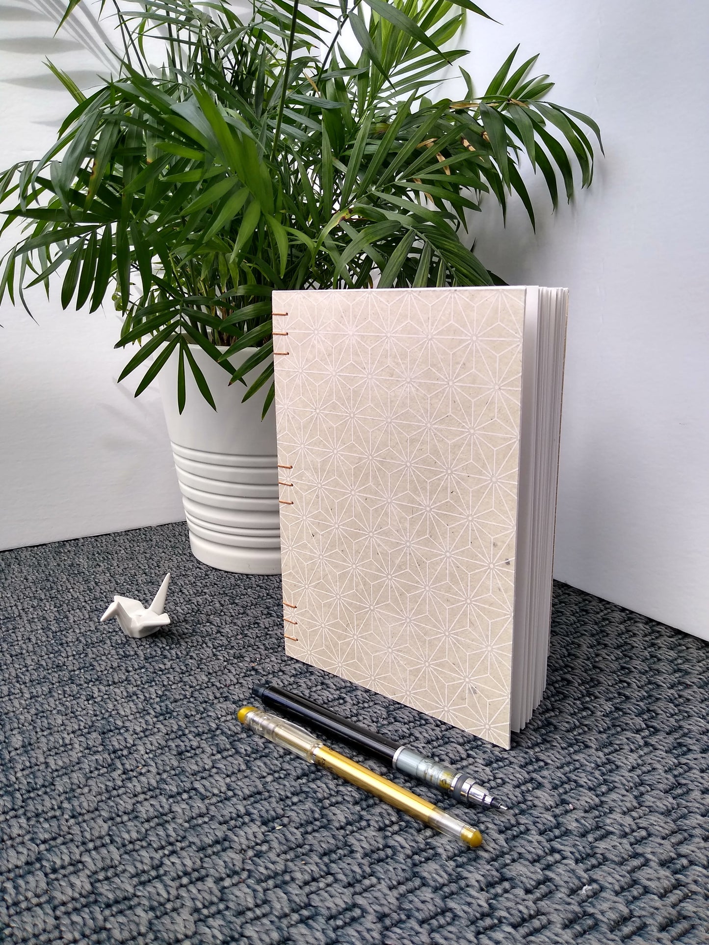 A handmade journal sits upright beside a potted plant, a ceramic crane, a gold pen, and a black mechanical pencil. The cover of the journal is cream with a white asanoha pattern on it. 