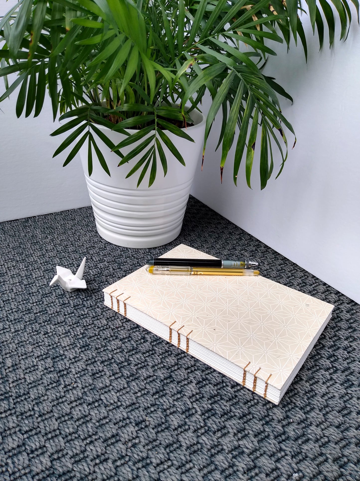 A handmade journal laying on a grey carpet beside a potted plant, and a ceramic crane. There is a gold pen and a black mechanical pencil on top of the journal. The cover of the journal is cream with a white asanoha pattern on it. The journal is turned to show the brown stitching along the spine, holding everything together.