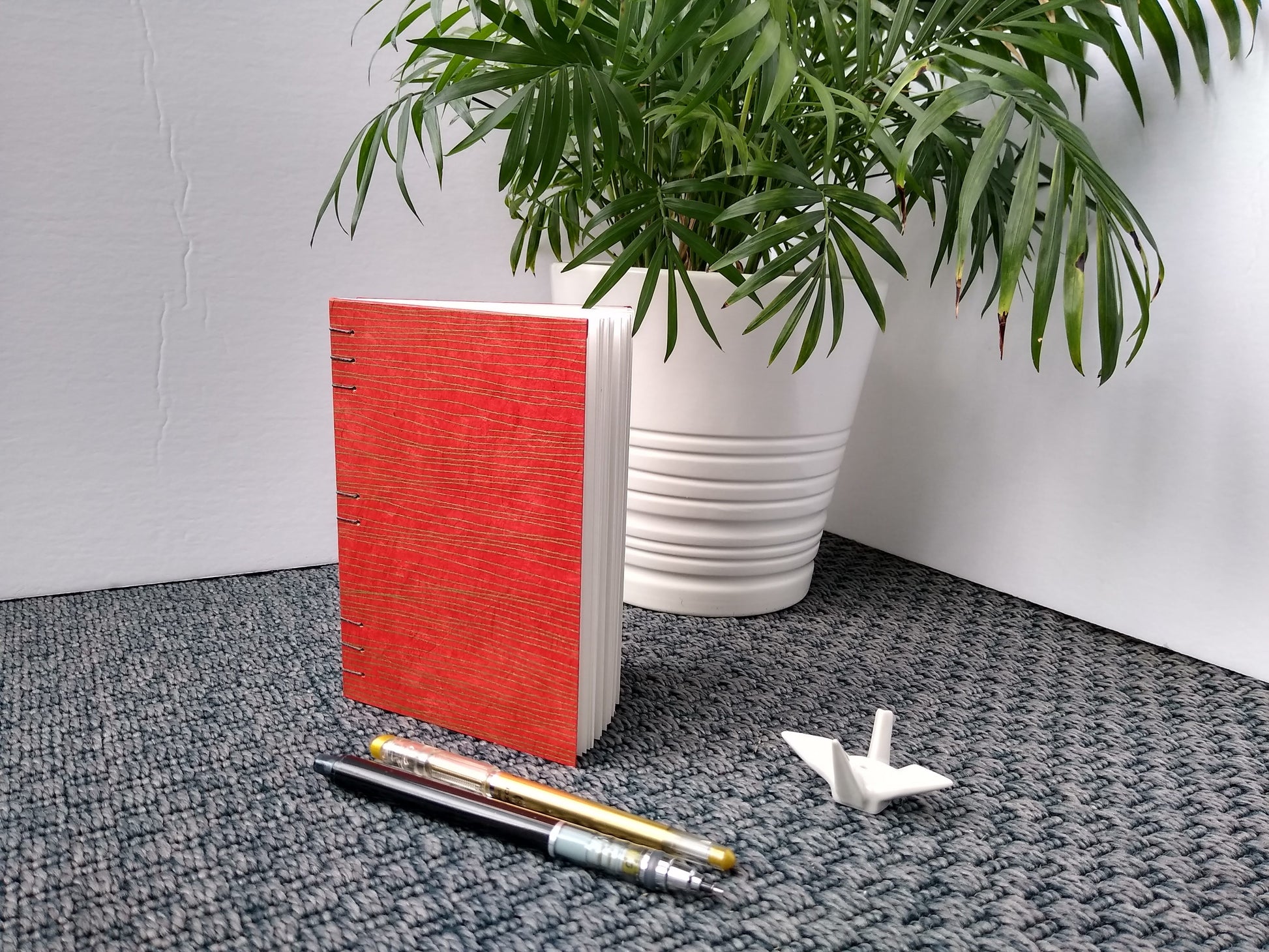 A handmade journal sits upright beside a potted plant, a ceramic crane, a gold pen, and a black mechanical pencil. The cover of the journal is red with wavy horizontal gold lines across it.