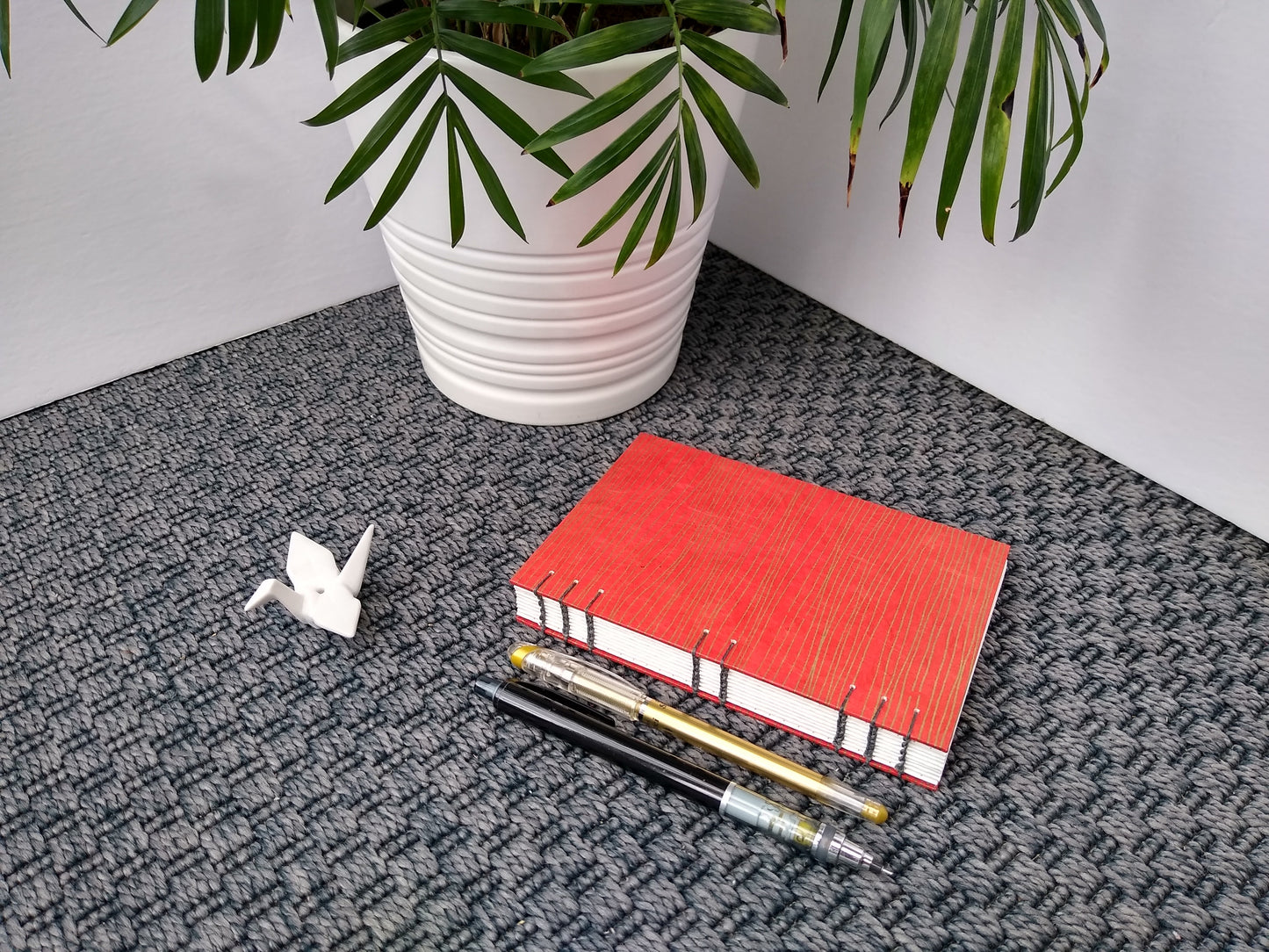 A handmade journal laying on a grey carpet beside a potted plant, a ceramic crane, a gold pen, and a black mechanical pencil. The cover of the journal is red with wavy horizontal gold lines. It is angled to show the open spine, stitched together with grey thread.