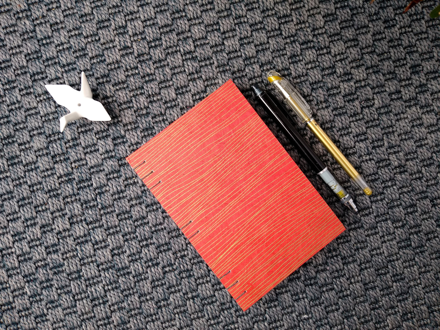 A handmade journal laying on a grey carpet beside a potted plant, a ceramic crane, a gold pen, and a black mechanical pencil. The cover of the journal is red with wavy horizontal gold lines.