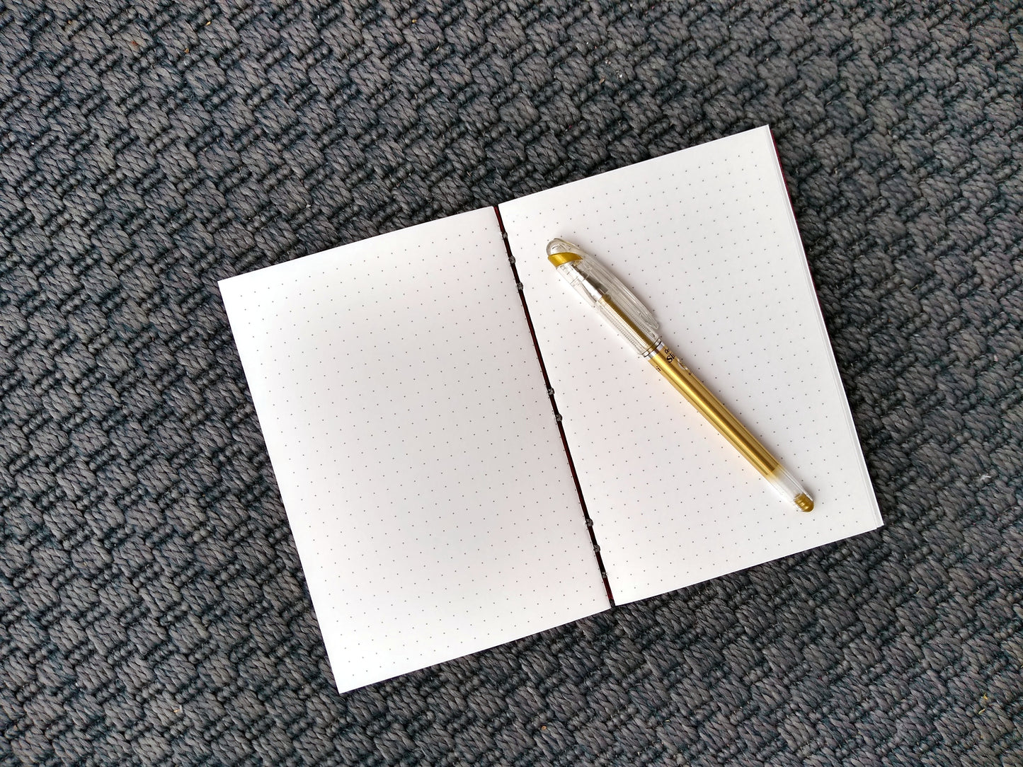 An open journal lays on a grey carpet. The journal has a grey thread connecting the pages at the spine and a gold pen rests across the white pages, printed with a dotted grid.