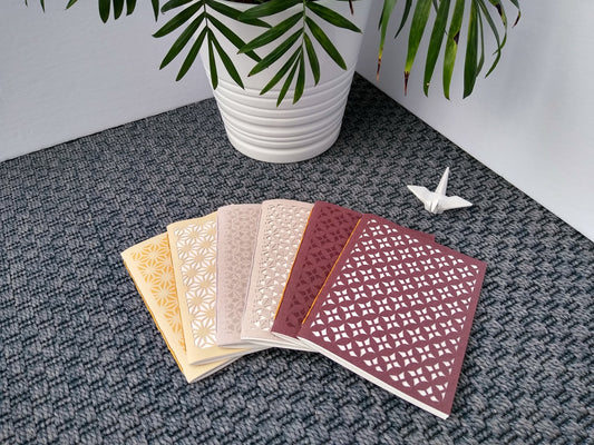Six handmade notebooks laying spread out on a grey carpet beside a potted plant and a ceramic crane. Two are cream colored, two are grey colored, two are burgundy colored. One of each color has a repeating pattern printed on it, the other of each color has the pattern cut out.