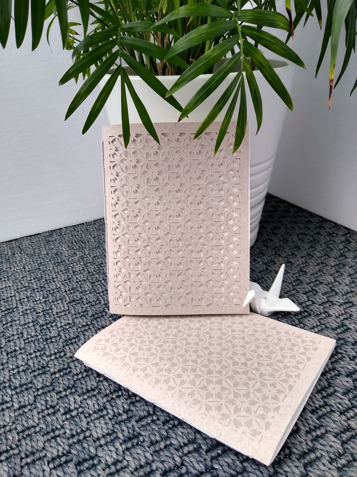 Two gray notebooks are displayed on a grey carpet. Next to them is a potted plant and ceramic crane. Both notebooks have a geometric and hexagonal floral pattern, repeated across the entire cover. One is cut the other is printed.