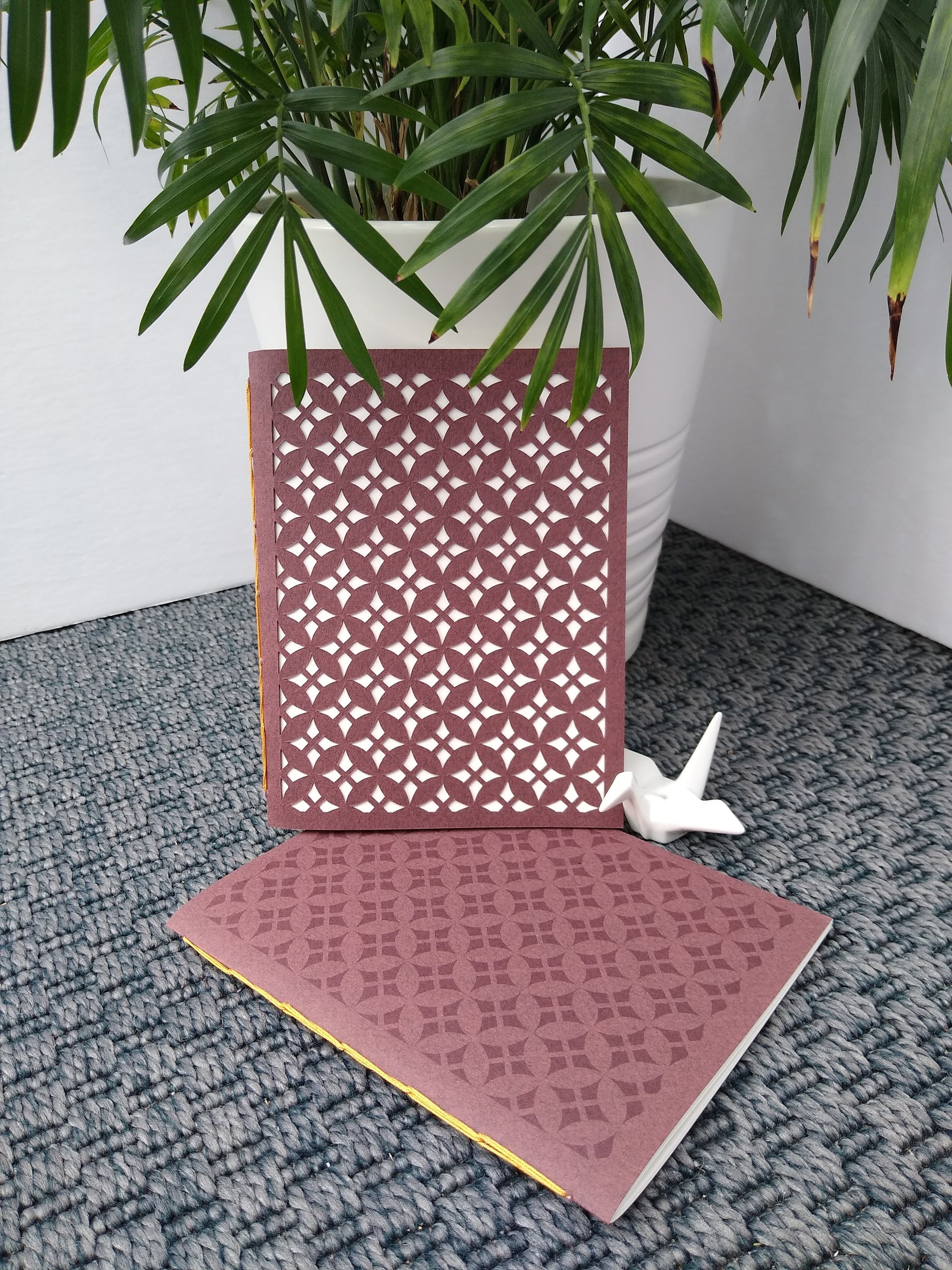 Two burgundy notebooks are displayed on a grey carpet. Next to them is a potted plant and ceramic crane. Both notebooks have a geometric diamond pattern repeated across the entire cover. One is cut the other is printed.