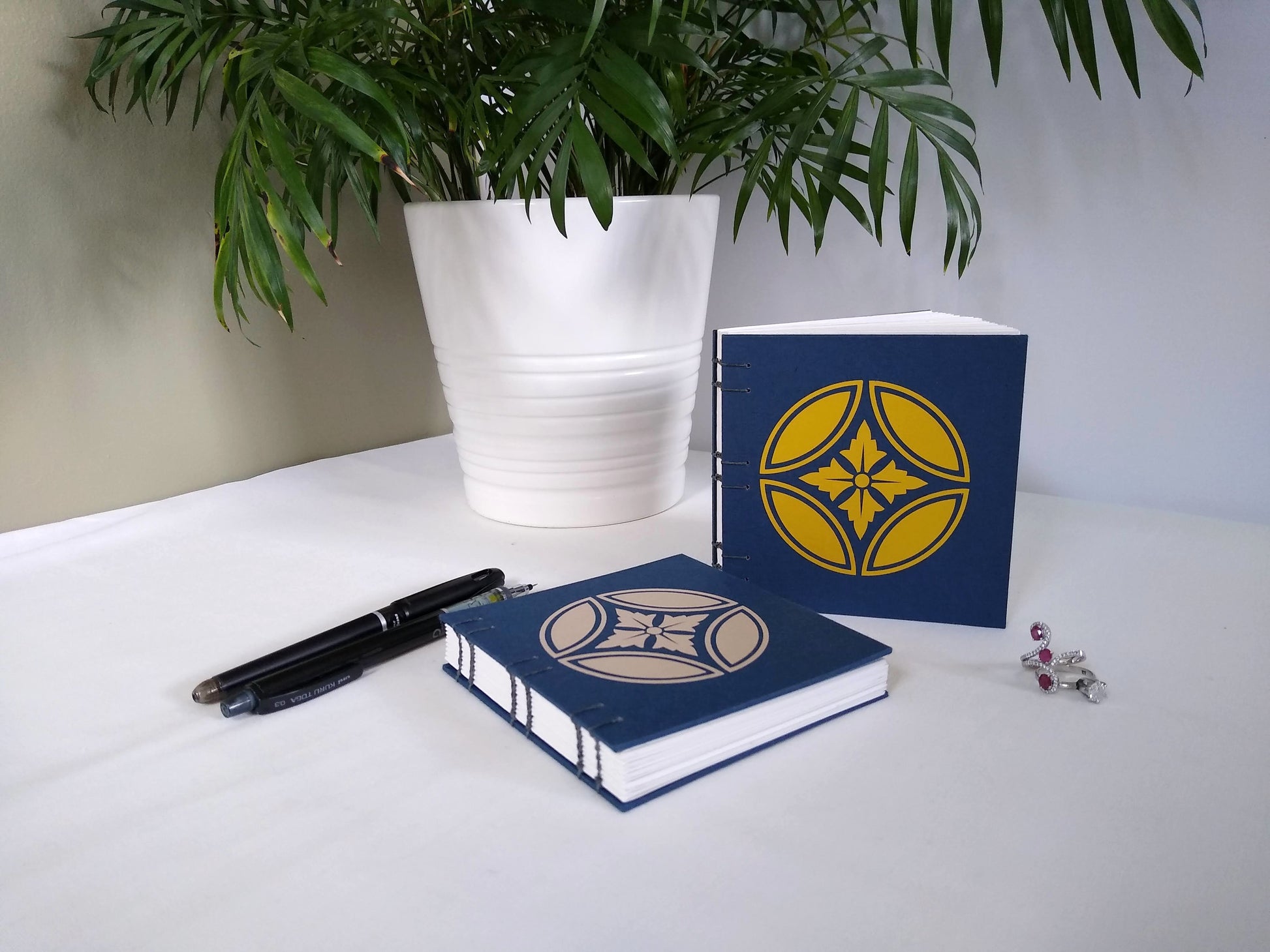 Two handmade journal are displayed on a desk, one sits upright beside a potted plant, the other lays flat. Two rings and a black pen and mechanical pencil are also on the desk. The cover of the journals are deep blue, with japanese inspired floral emblems appliqued on them. The one on the upright journal is in gold, the other is taupe.