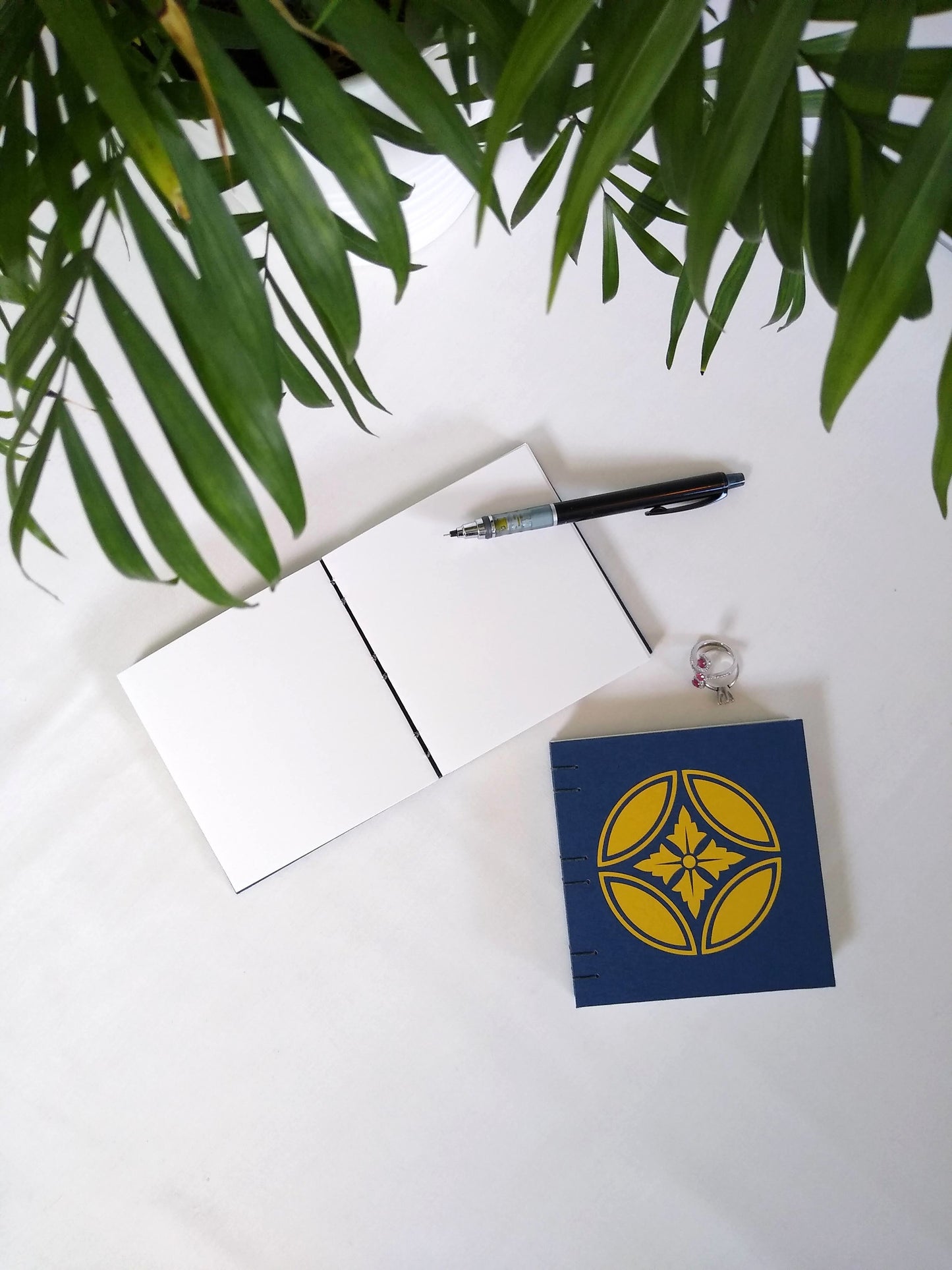 An open journal lays on a white desk next to a potted plant. The journal has a blue thread connecting the pages at the spine and a black mechanical pencil rests across the blank white pages. Beside it are two rings and another journal, closed, with a gold japanese inspired floral emblem on the cover.