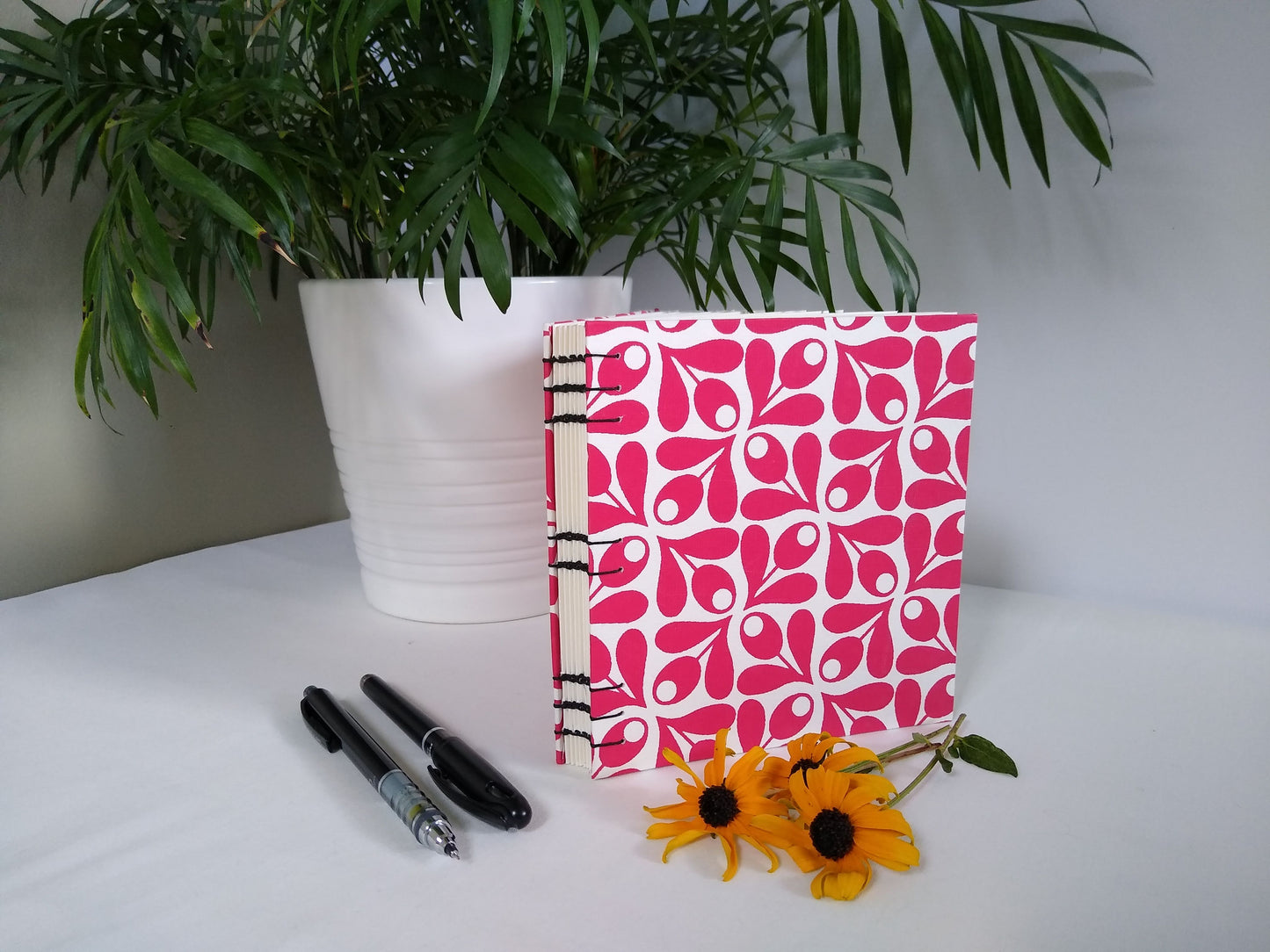 A handmade journal sits upright beside a potted plant, three yellow flowers, a black pen, and a black mechanical pencil. The cover of the journal is cream with red olive springs repeated across it. It's angled so the open spine is visible, along with the black stitching holding it together.