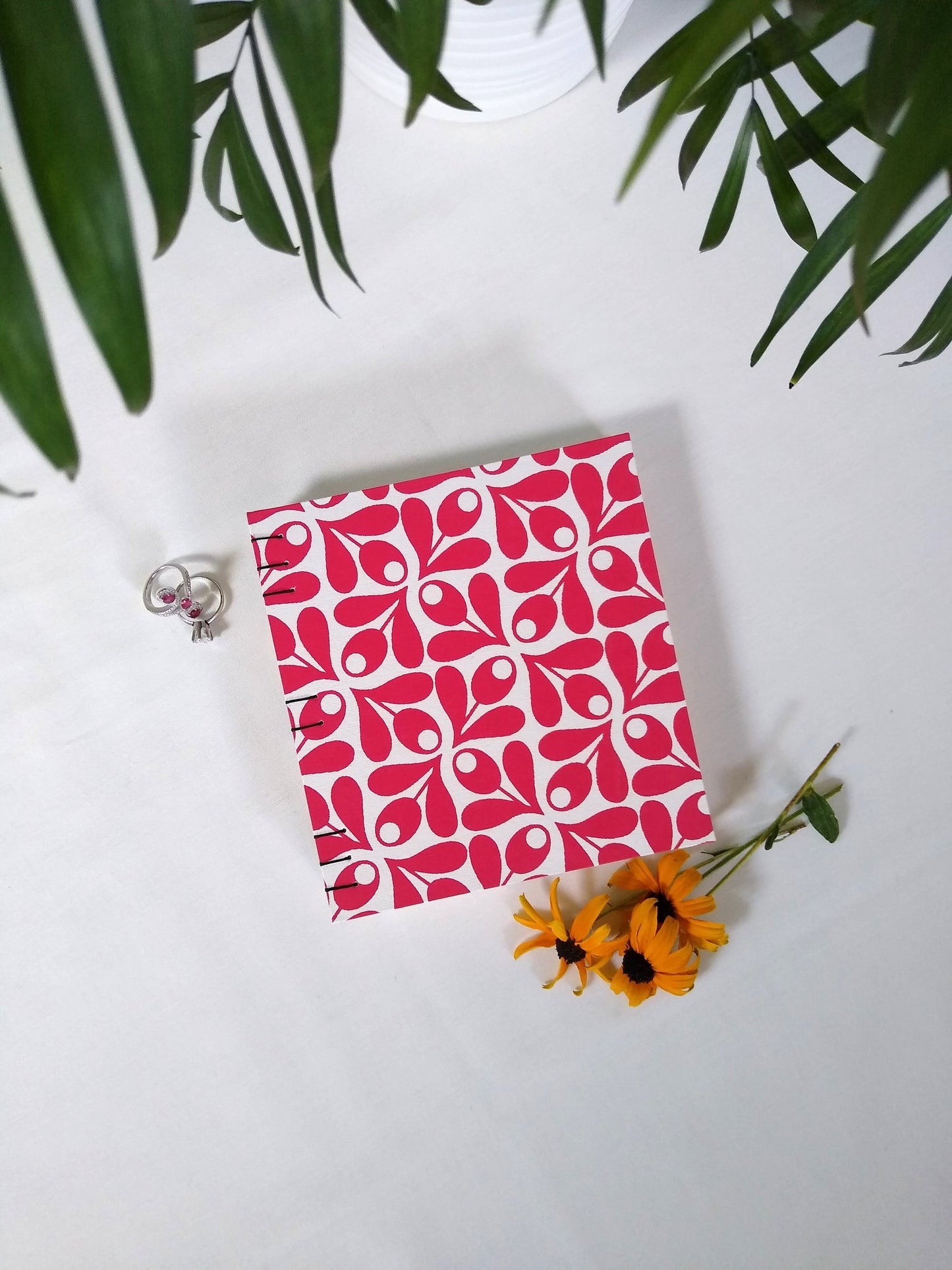 A handmade journal laying on a white desk beside a potted plant, two rings, and three yellow flowers. The cover of the journal is cream with red olive springs repeated across it.