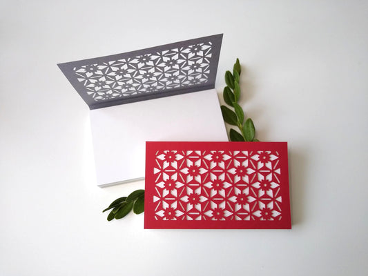 Two small notepads rest on a white background. One, with a gray cover, is open. The other, with a red cover, is closed. There are two sprigs of green leaves next to them. Cut into the covers is a geometric and hexagonal floral pattern, repeated across the entire cover.