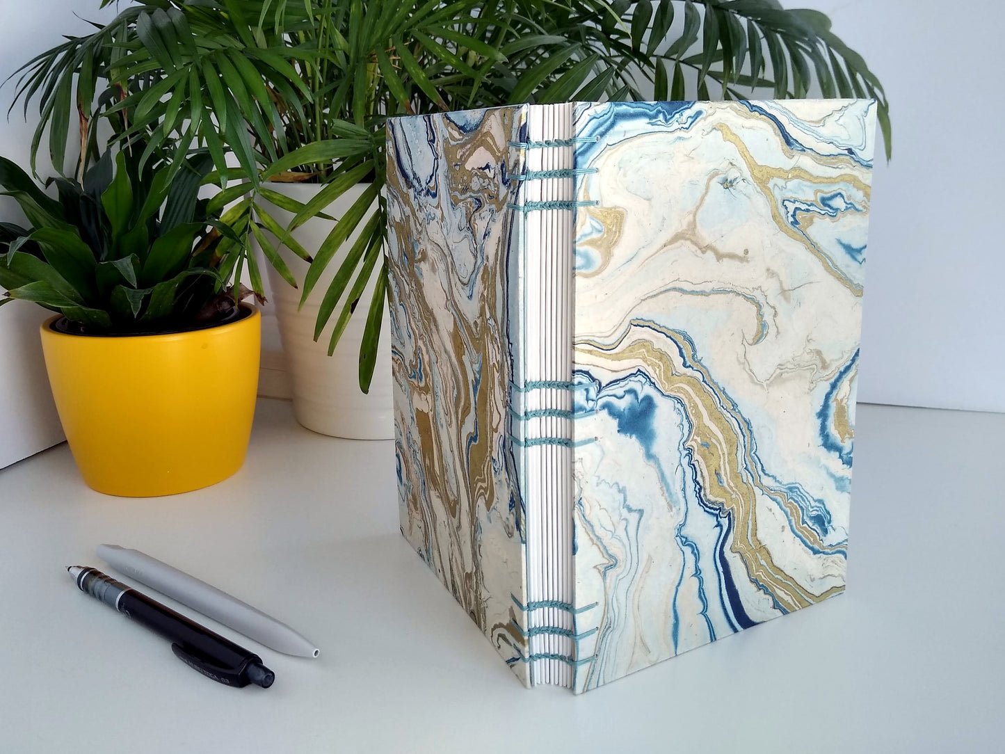 A handmade journal sits upright beside two potted plants, a grey pen, and a black mechanical pencil. The cover of the journal is cream paper, marbled with blue and gold. It's turned to show the open spine and the blue stitching holding it together.