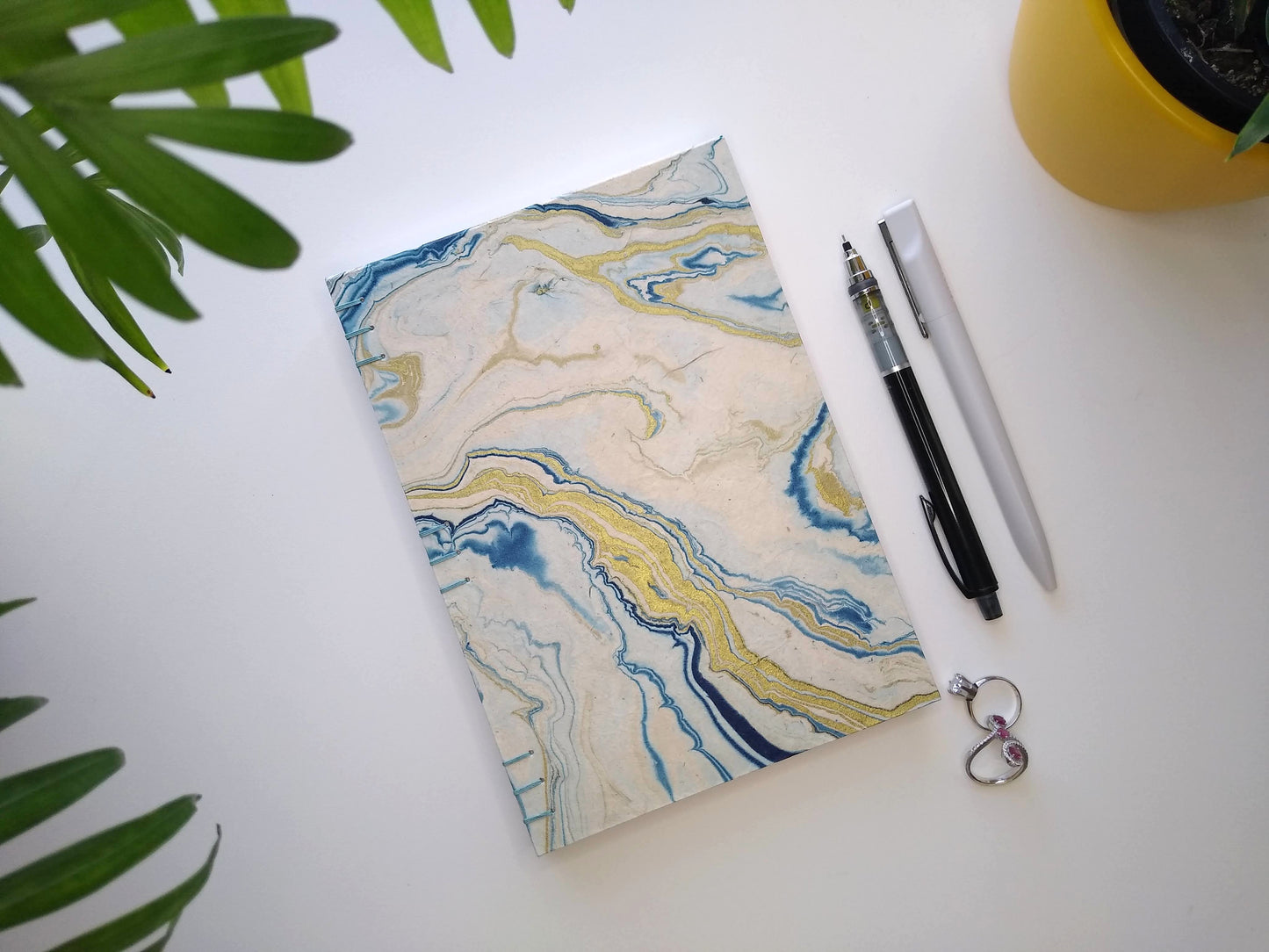 A handmade journal laying on a white desk beside two potted plants, a pair of rings, a grey pen, and a black mechanical pencil. The cover of the journal is cream paper, marbled with blue and gold.