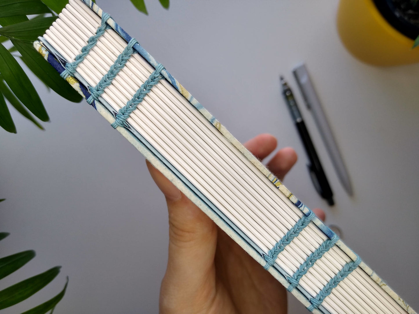 A hand holds the journal up, showing a close up of the open spine and the blue thread used to stitch it. There are two sets of three columns of stitches visible. A white desk, two potted plants, a black mechanical pencil and a grey pen are out of focus in the background.