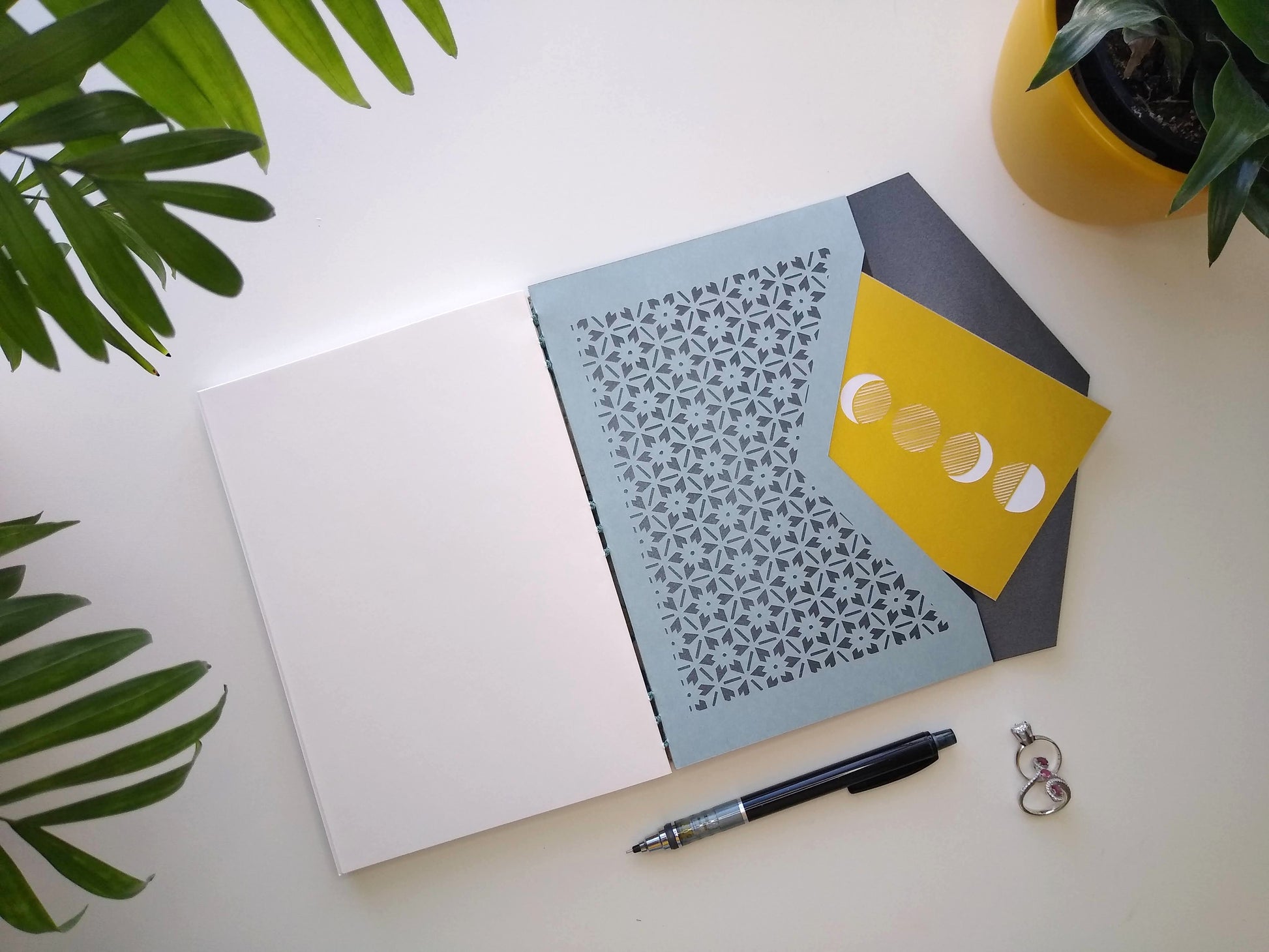 An open journal lays on a white desk next to two potted plants, a pair of rings, and a black mechanical pencil. The journal shows blank white pages on the left side and a blue envelope on the right. The envelope has a floral lattice design cut into it, lined with metallic grey paper. It is open and has a gold, moon phases postcard sticking out of it.