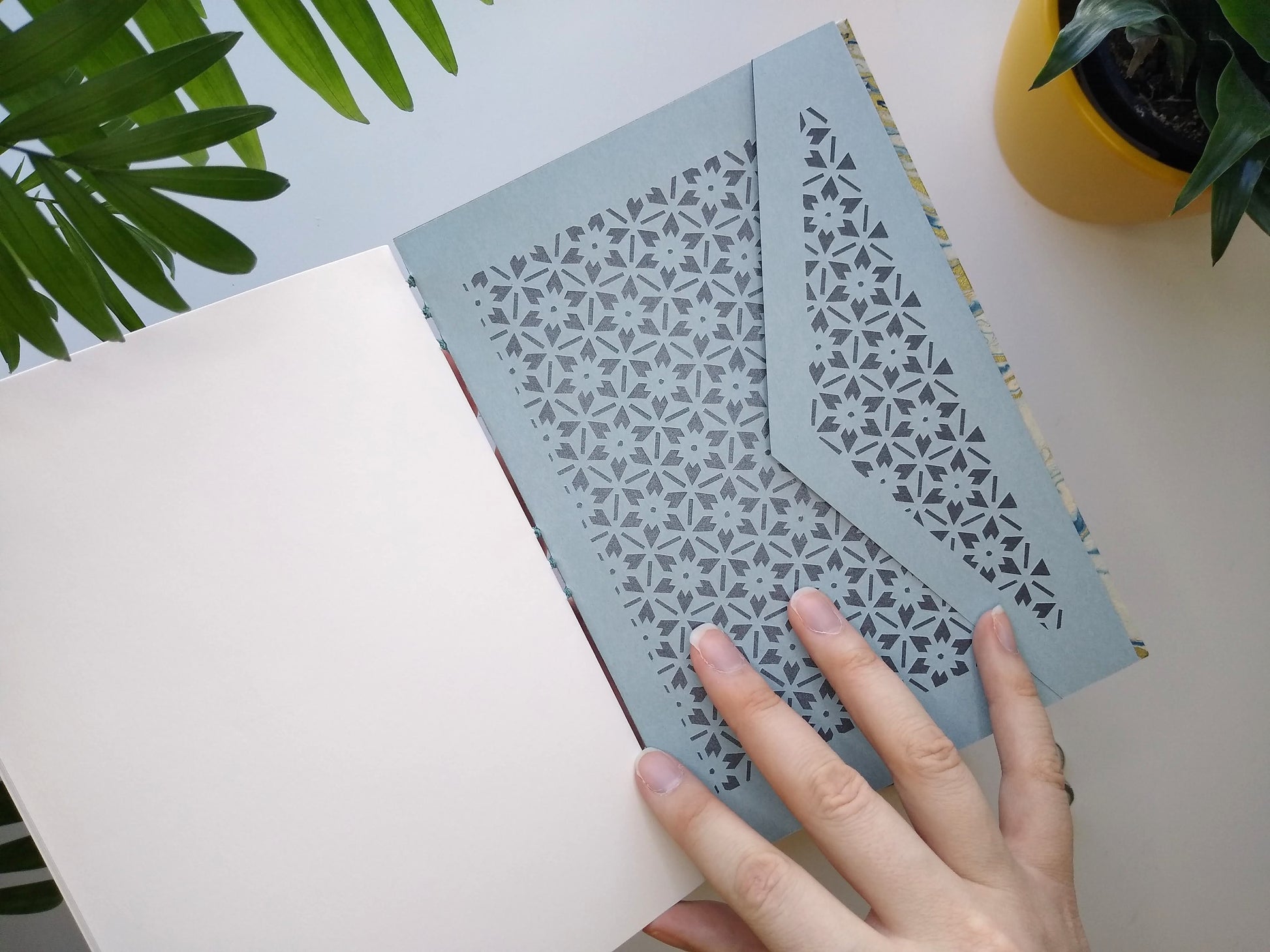 A journal is held open by a hand, showing a blue envelope at the back. The envelope has a floral lattice design cut into it, backed with metallic grey paper. It's held over a white desk with two potted plants in view.