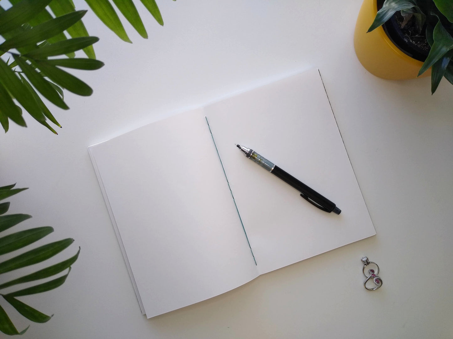 An open journal lays on a white desk next to two potted plants and a pair of rings. The journal has a blue thread connecting the pages at the spine and a black mechanical pencil rests across the blank white pages.