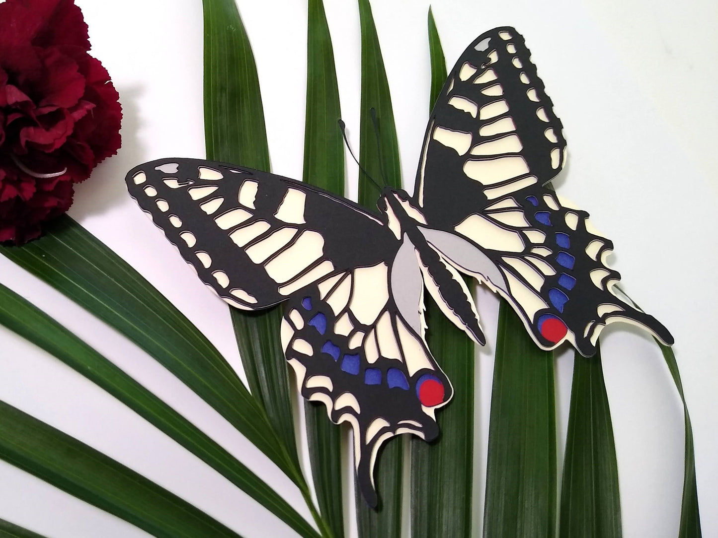 Multi-layered paper butterfly, in the design of a Old World Swallowtail butterfly. It rests on top of a sprig of leaves and beside a dark red flower.